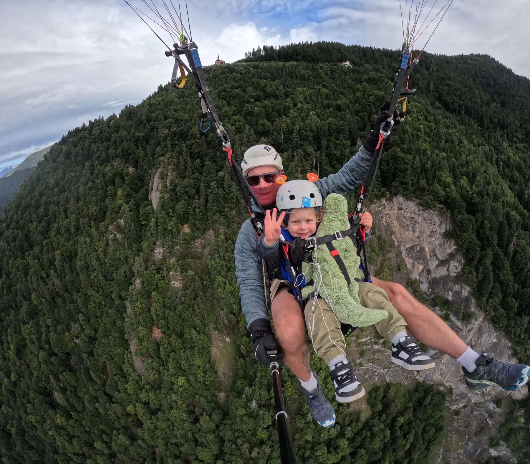 Pilot and young passenger waving mid-flight over forested slopes near Interlaken
